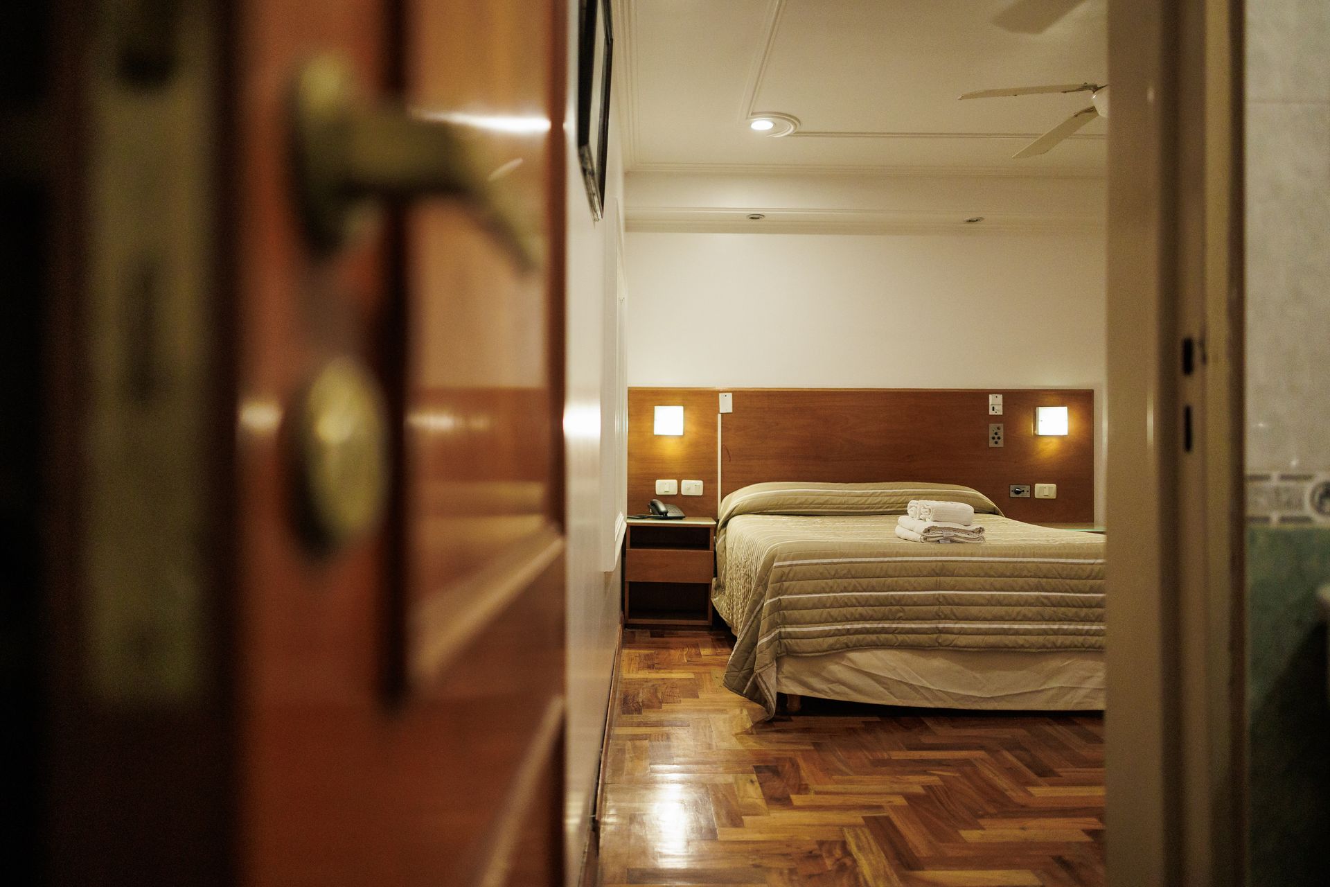 Interior del dormitorio visto a través de una puerta de madera abierta. Cama, mesita de noche y suelo de madera.