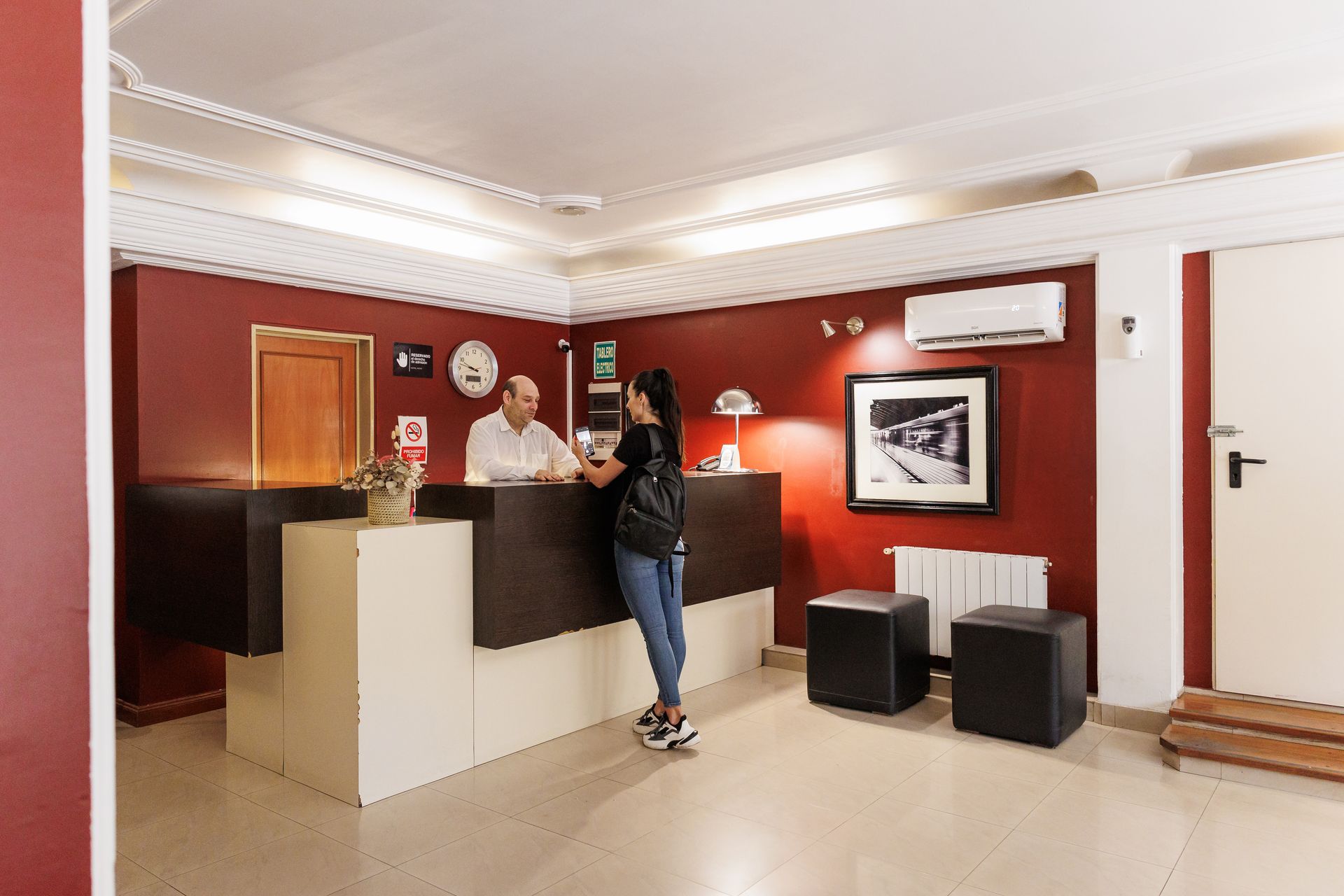 Vestíbulo de hotel con un huésped en recepción registrándose. Un hombre le asiste. Paredes y decoración rojas.