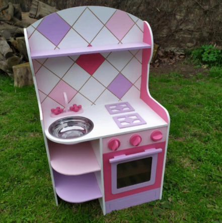 Una cocina infantil de madera, pintada de rosa, morado y blanco, sobre césped. Incluye fregadero, estufa y horno.