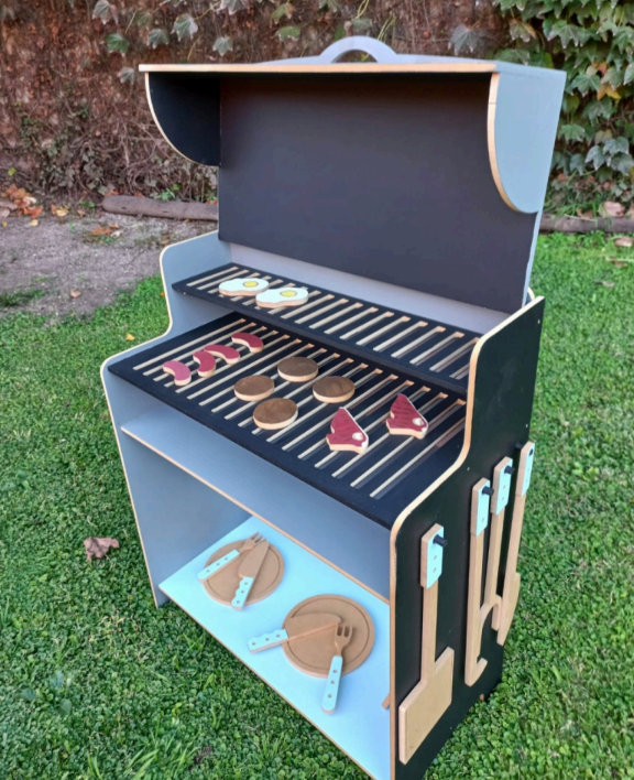 Parrilla de juguete de madera con comida y utensilios sobre césped verde.