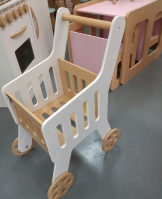 Carrito de compras de juguete de madera, marco blanco, interior y ruedas de color beige, sentado sobre una superficie gris.