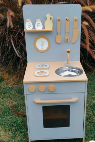 Cocina de juguete de madera en color gris y tostado con utensilios, fregadero y horno en un entorno exterior.