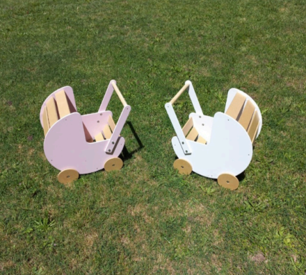 Dos cochecitos de bebé de juguete de madera, uno rosa y otro blanco, sobre césped verde.
