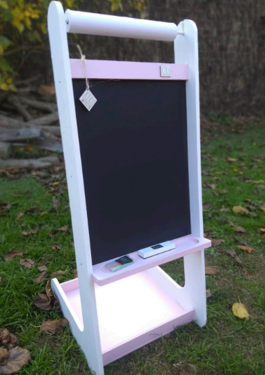 Caballete infantil de madera de color rosa y blanco con pizarra, colocado al aire libre sobre el césped.