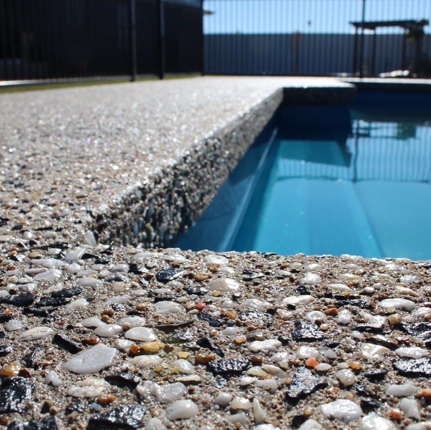 Close-up of a concrete pool deck with exposed aggregate stones. The pool's blue water reflects.