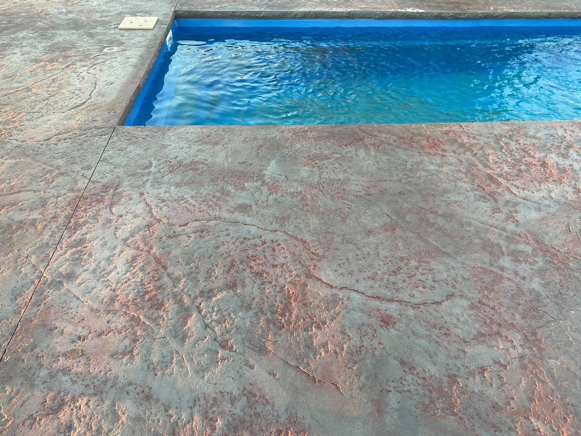 Concrete pool deck with reddish-brown and gray coloring, bordering a blue swimming pool.