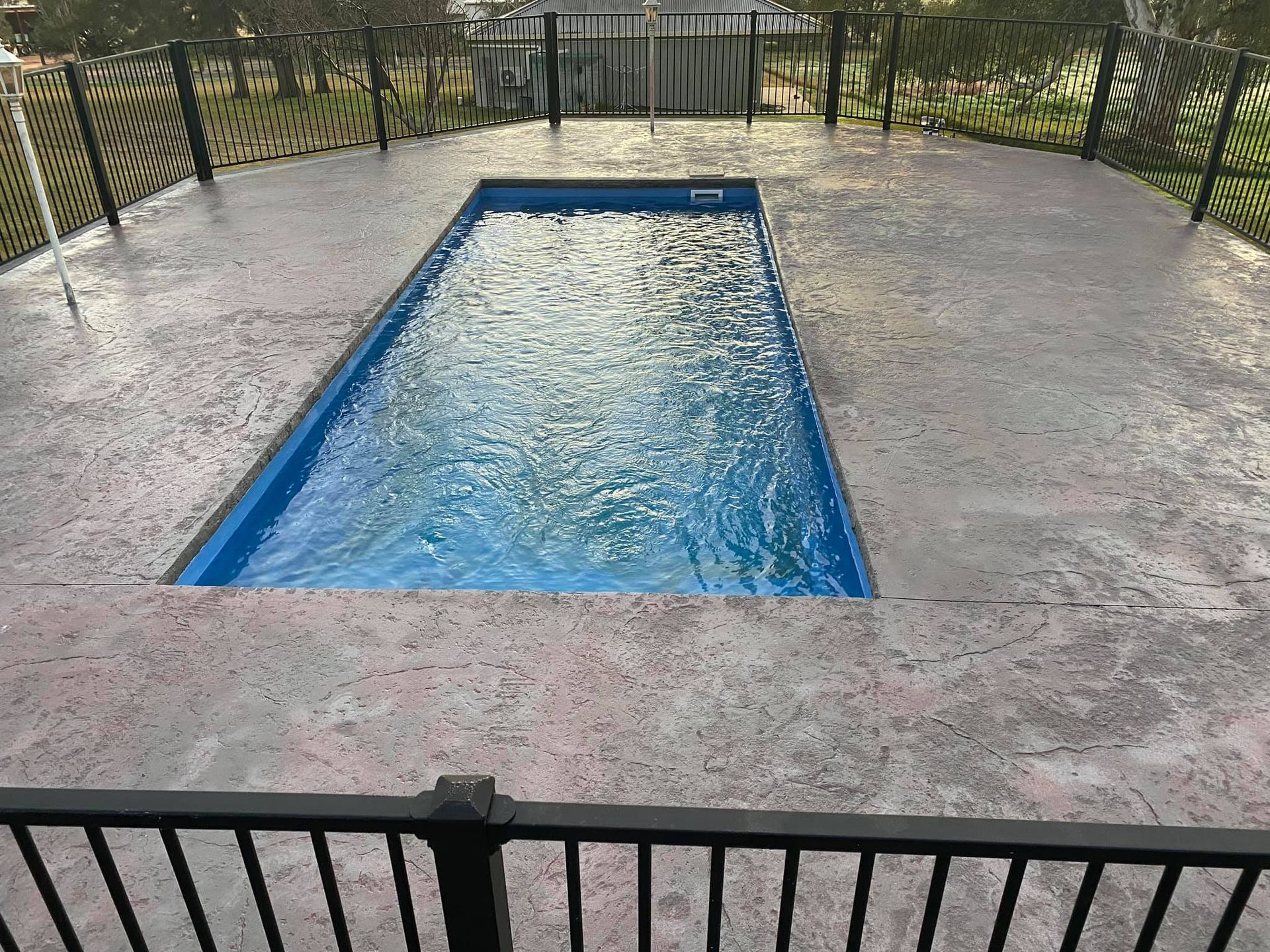 Rectangular blue swimming pool surrounded by gray concrete patio and black metal fence.