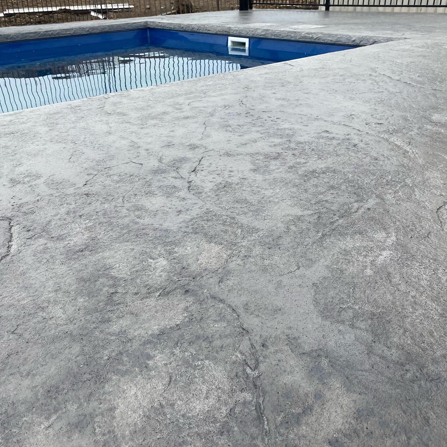 Concrete pool deck next to a blue pool; gray and textured.