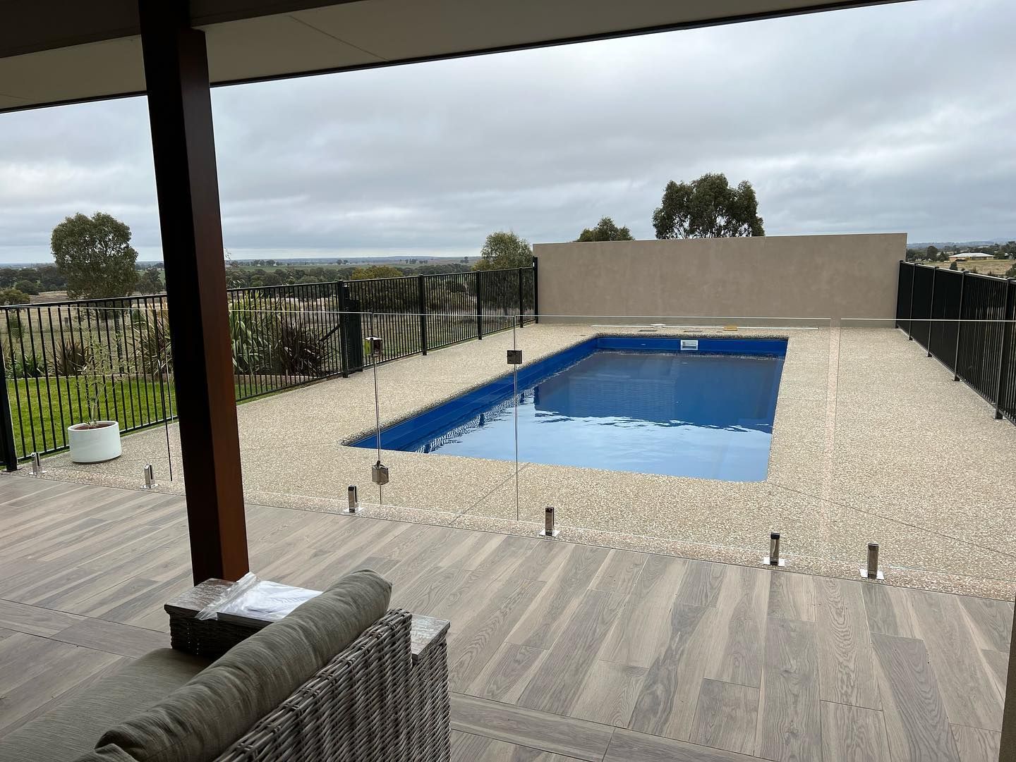 Poolside view: rectangular blue pool, light stone surround, gray deck, with a distant landscape under an overcast sky.