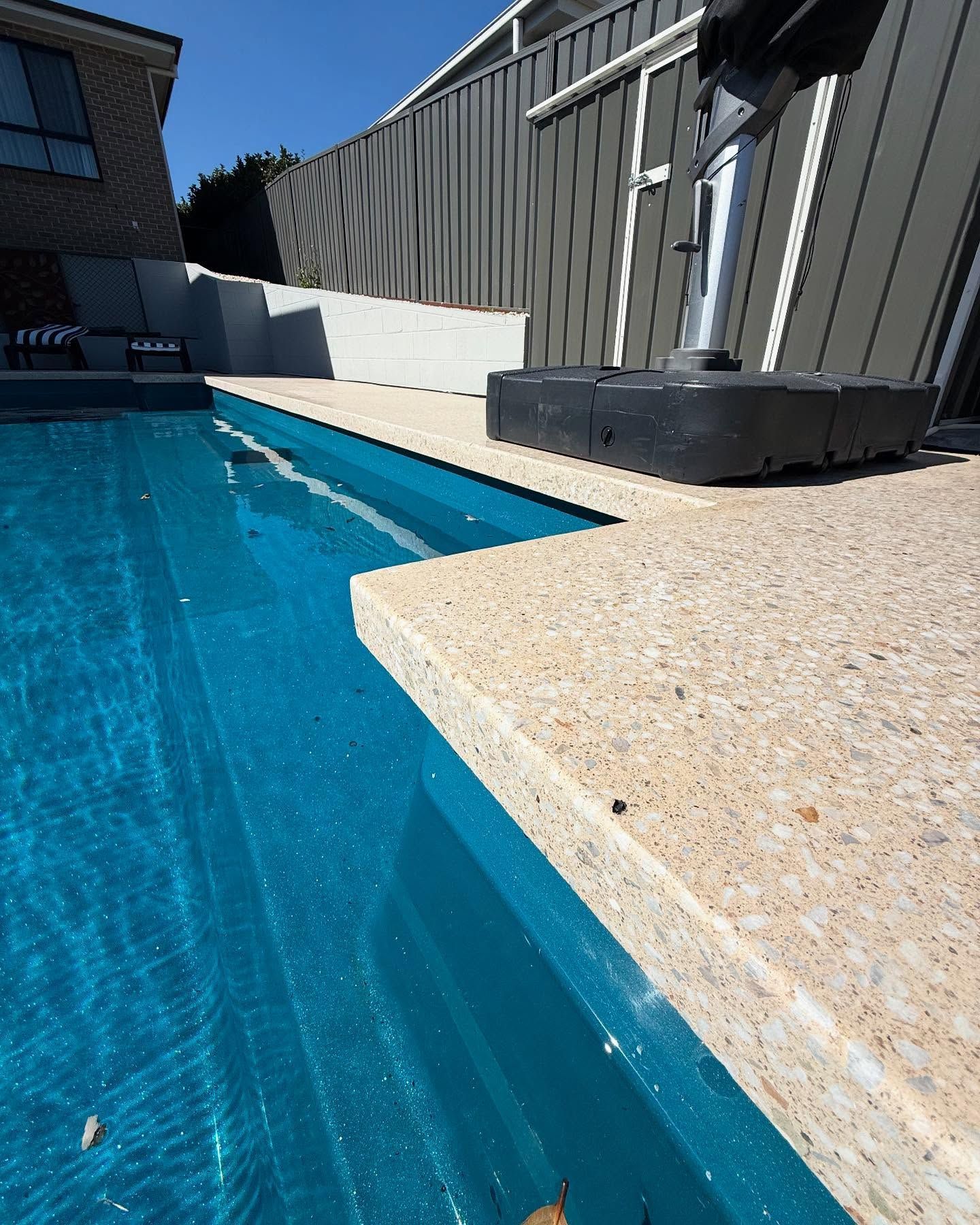 Swimming pool with turquoise water and beige concrete edging. Grey fence and blue sky in background.
