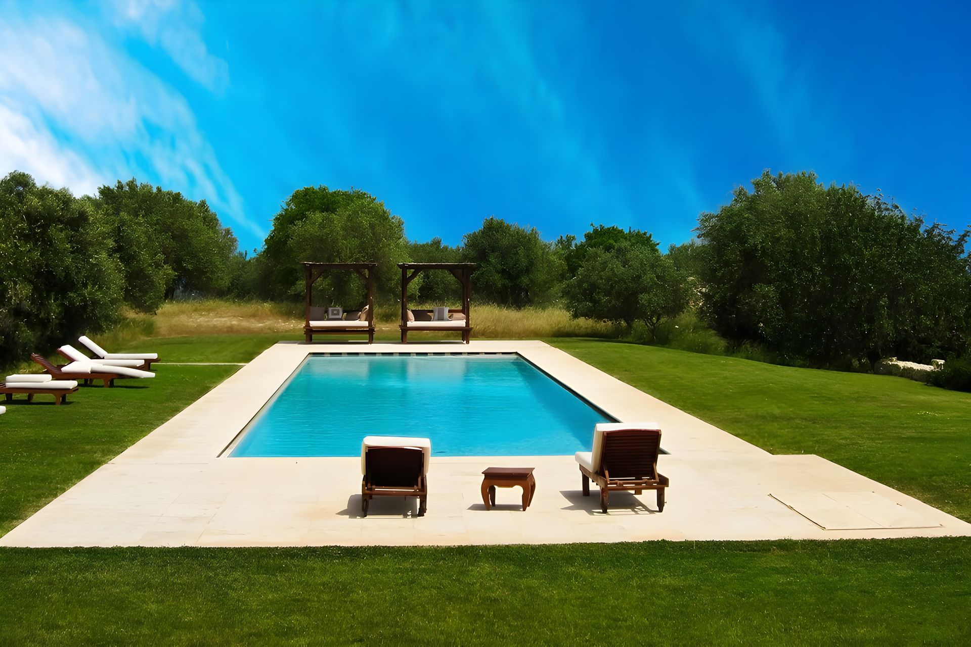 Swimming pool surrounded by green grass and trees under a blue sky. Two lounge chairs face the water.