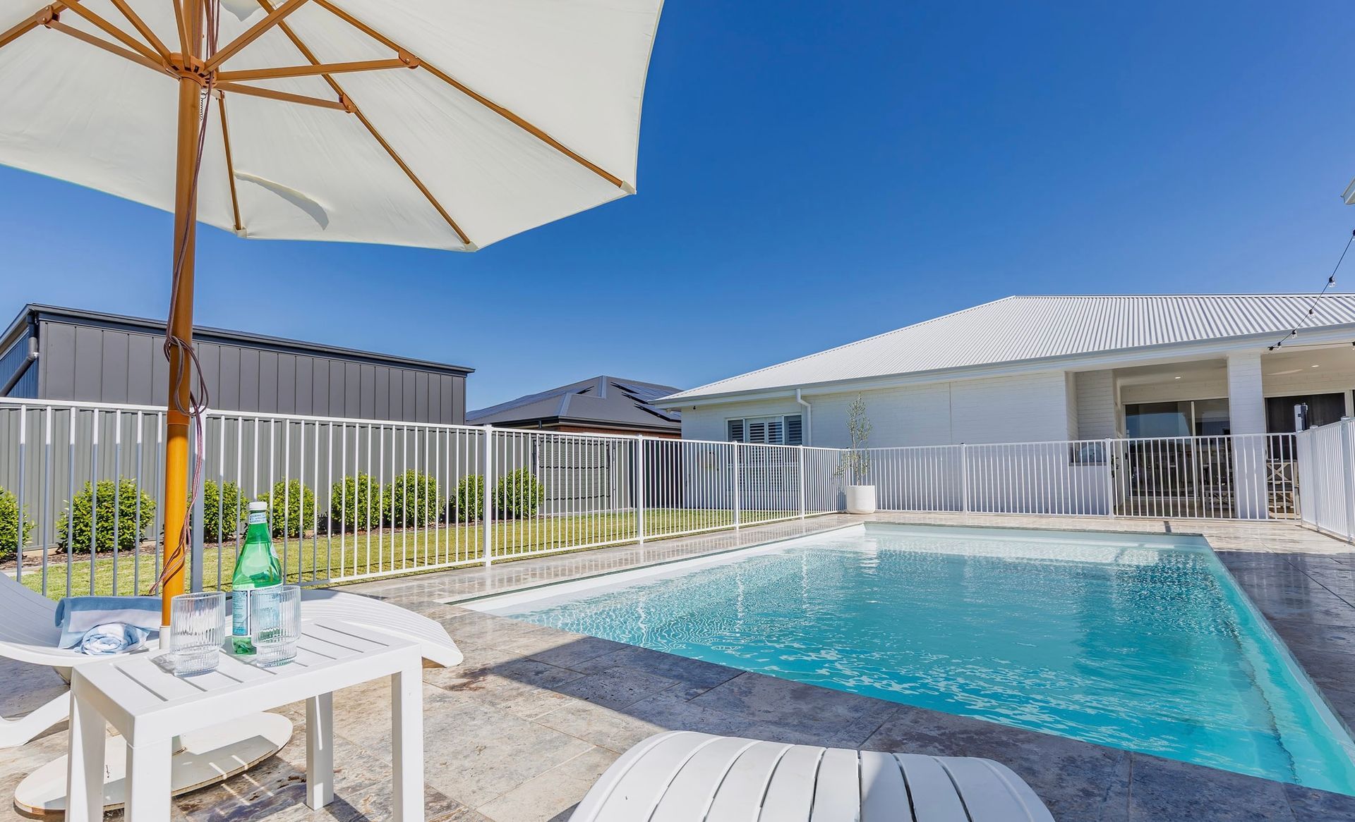 Poolside scene: White house, umbrella, pool, white fence, water bottle on a table, blue sky.