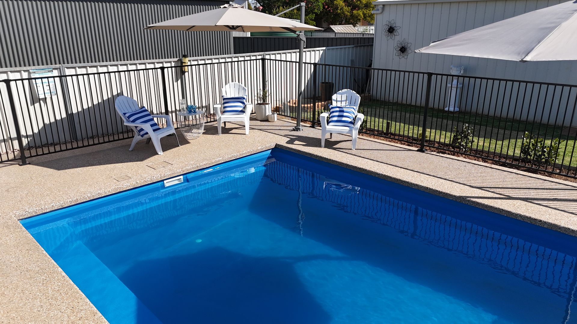 Pool with blue water, surrounded by a paved patio with white chairs, a black fence, and umbrellas for shade.