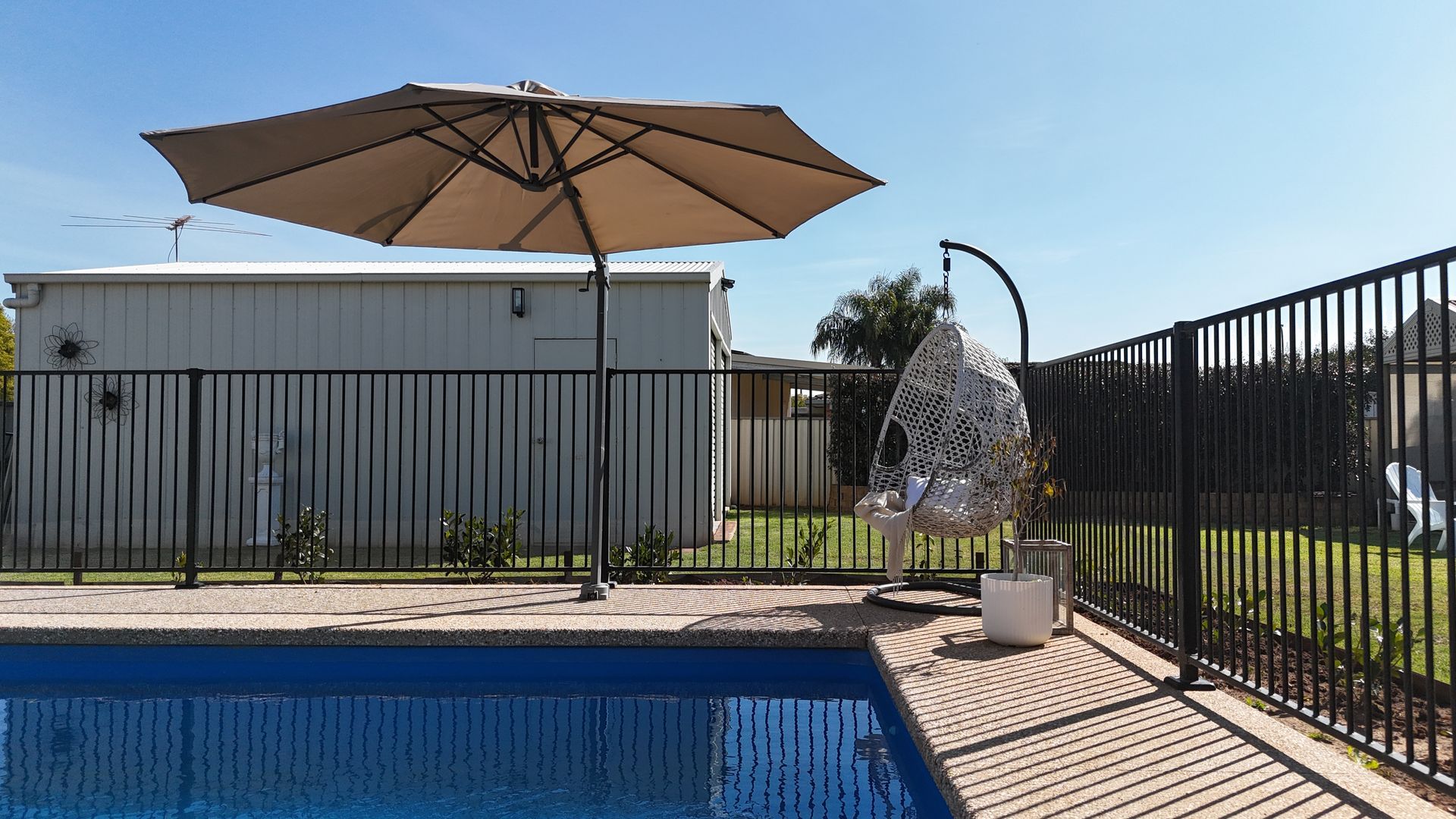 Poolside scene with a beige umbrella, hanging chair, and black fence against a bright blue sky.