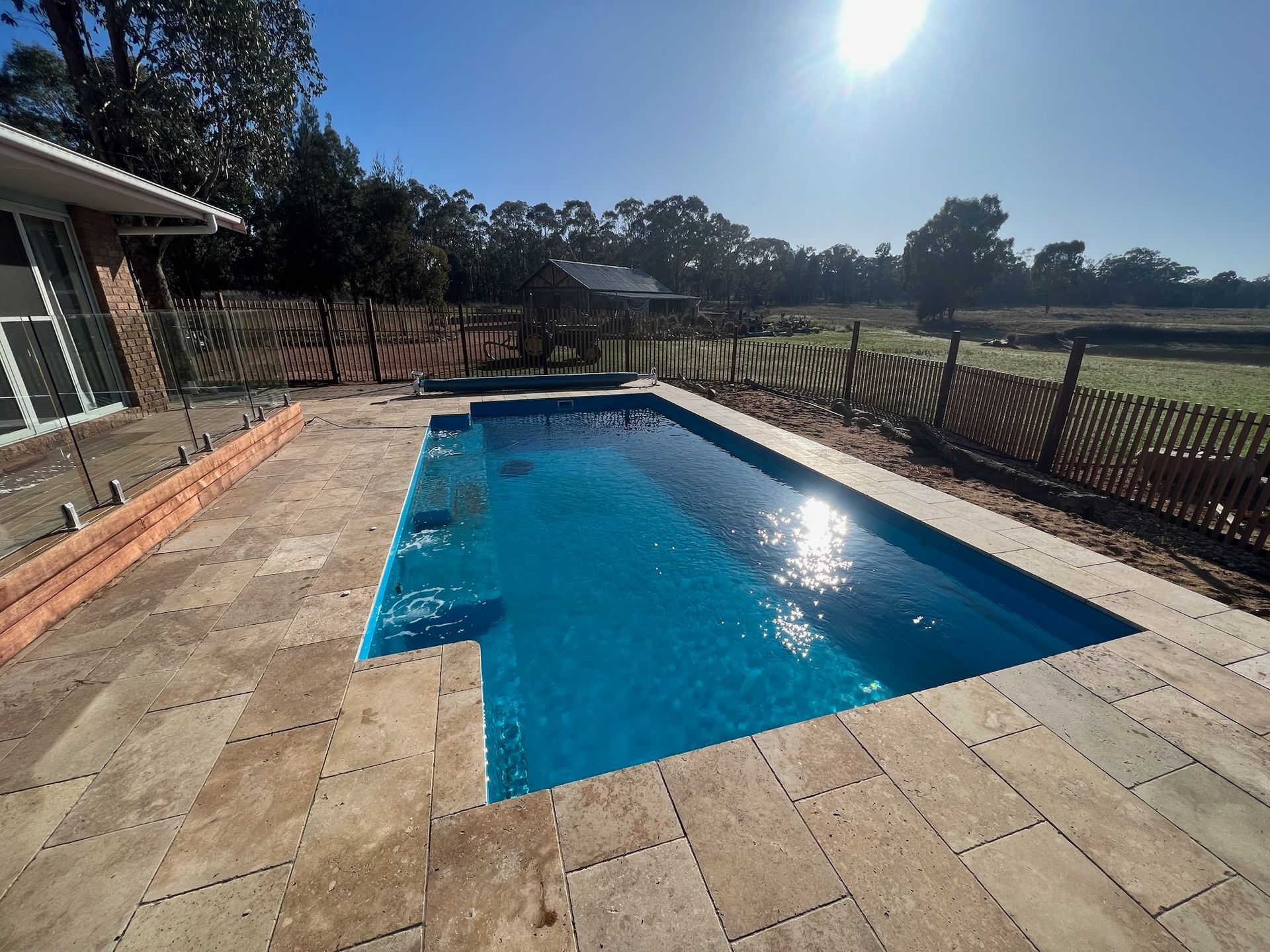 Rectangular blue pool with stone patio next to a house, sunny outdoor setting.