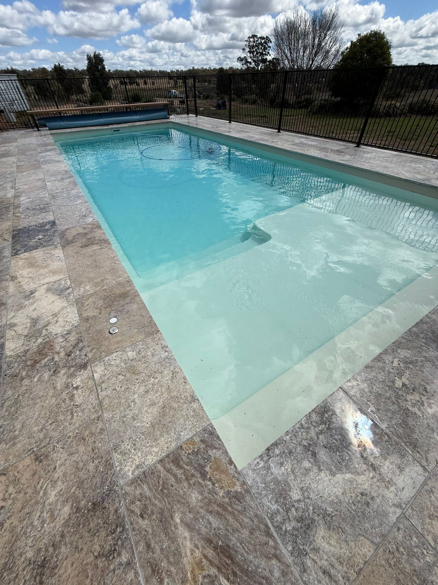 Rectangular swimming pool with light blue water and stone patio, set outdoors under a cloudy sky.