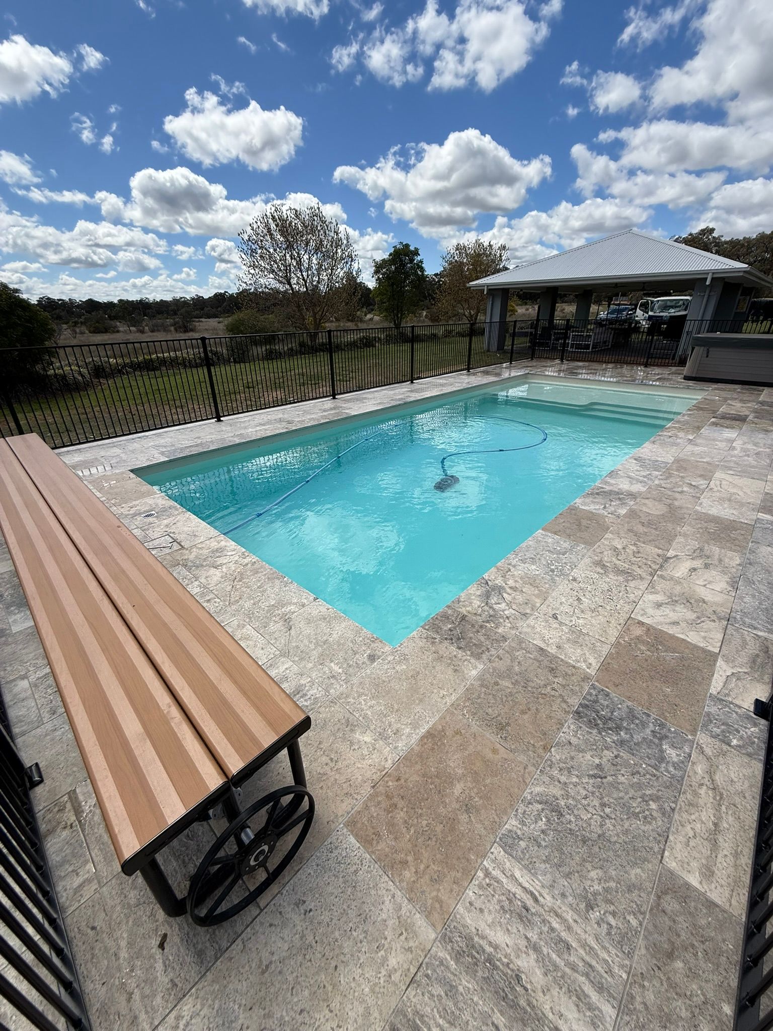 Swimming pool with a bench, stone patio, and gazebo on a sunny day with a blue sky.
