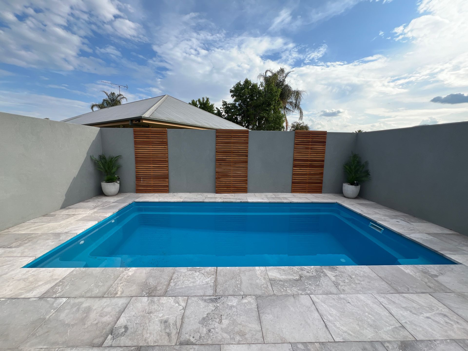 A rectangular blue swimming pool surrounded by gray stone. Wooden panels adorn a gray wall under a blue sky.
