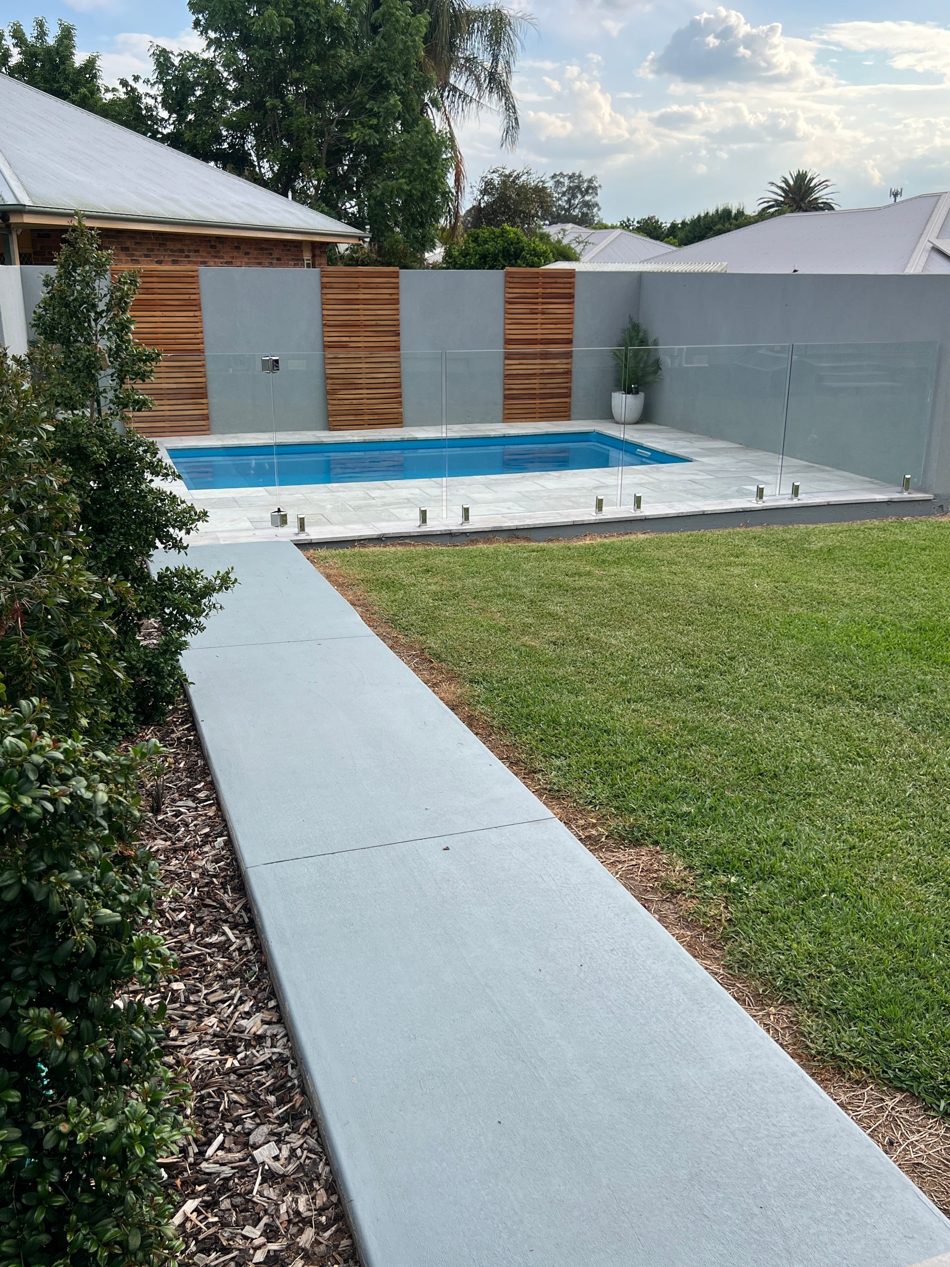 Backyard with pool, concrete pathway, wooden fence, and green lawn on a sunny day.