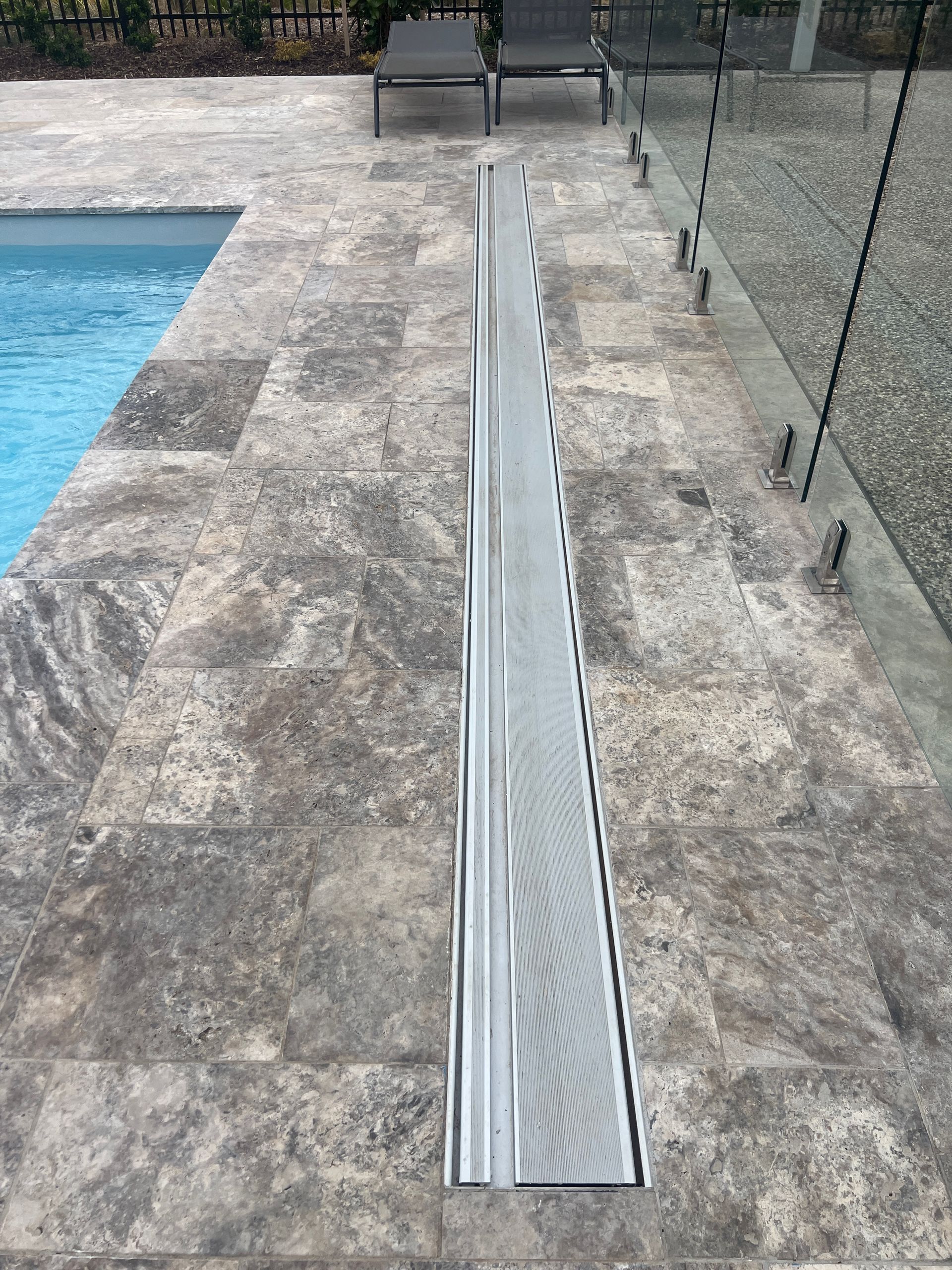 Poolside view with a long, metallic grate built into the stone flooring. Glass fence and pool visible.