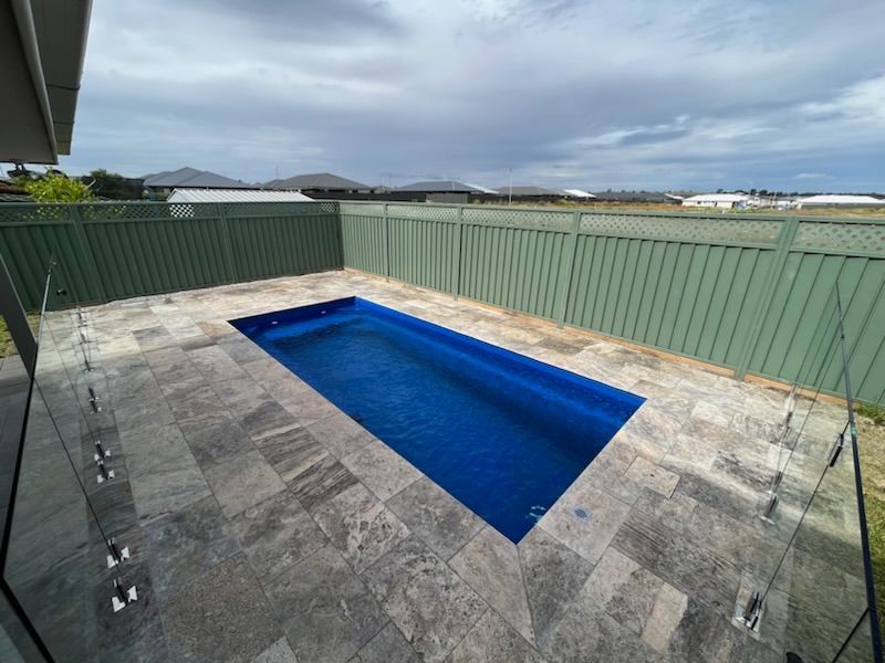 Rectangular blue pool in a paved backyard, surrounded by a green fence and cloudy sky.