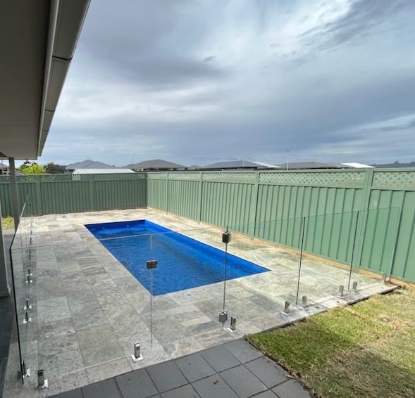 Backyard pool with glass fencing, gray stone, green fence, and cloudy sky.