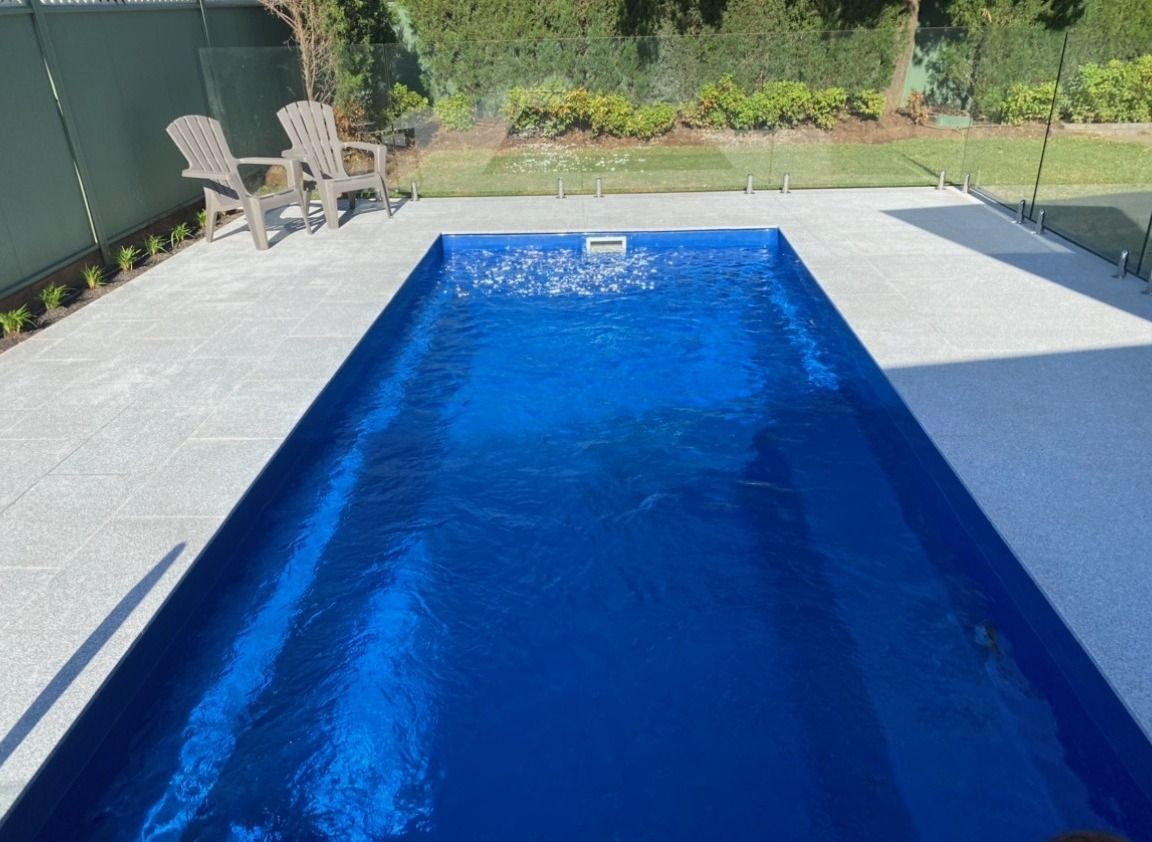 Rectangular blue swimming pool, light grey concrete patio, chairs, and glass fence. Green lawn and hedge in background.
