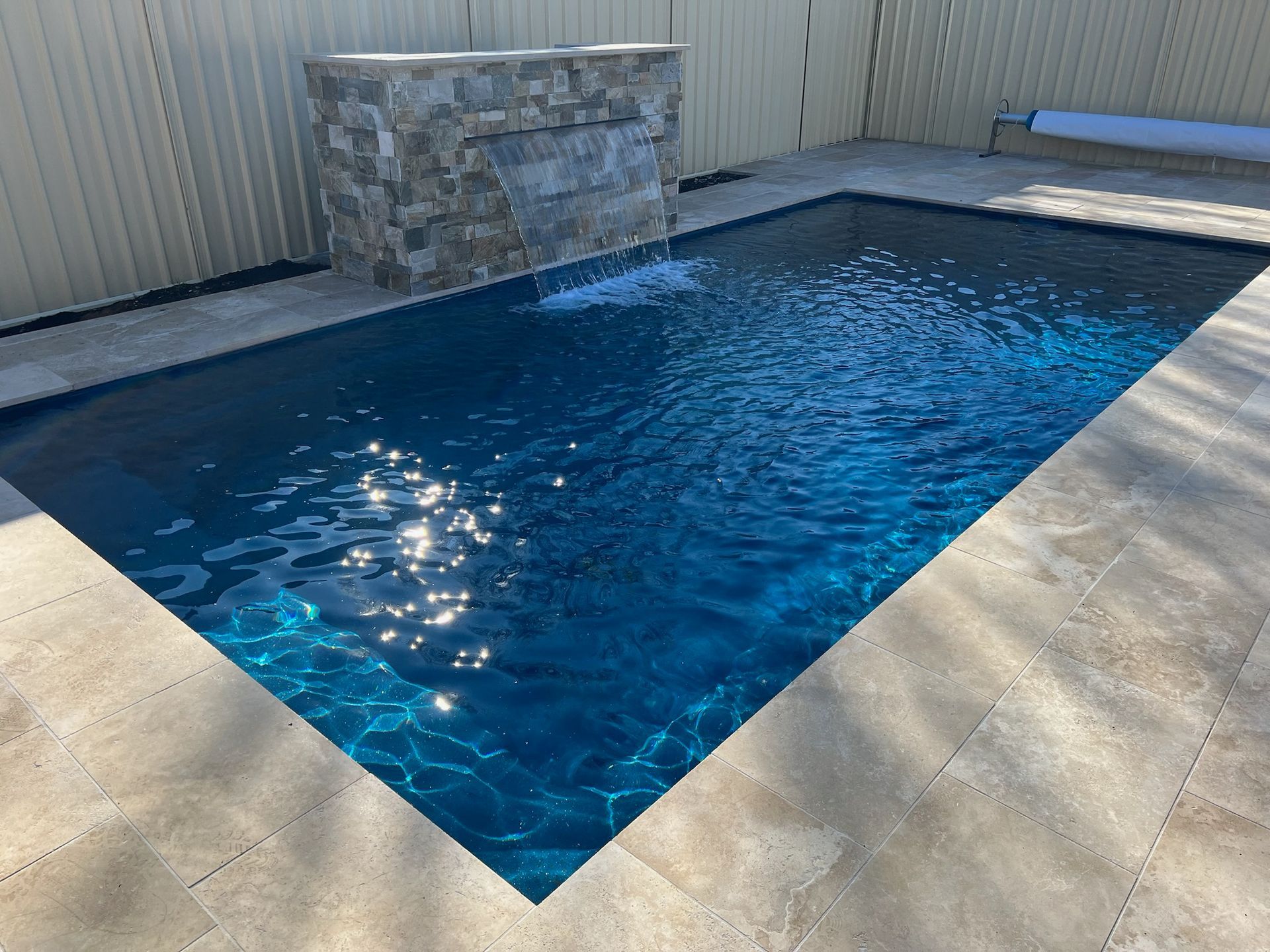 Rectangular swimming pool with blue water, stone waterfall feature, and light-colored paving.