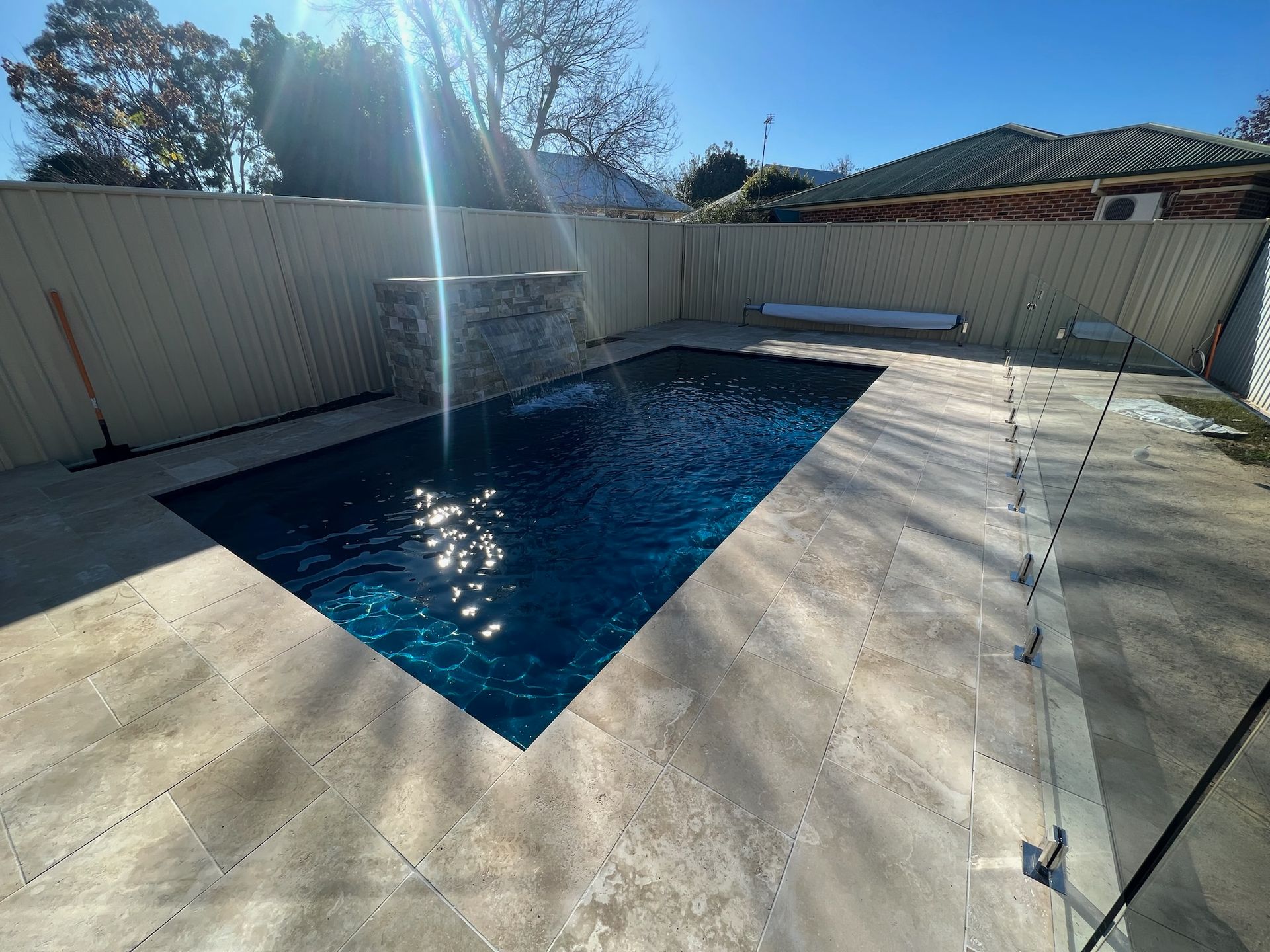 A rectangular swimming pool surrounded by beige stone and a light-colored fence. Bright sunlight reflects off the water.
