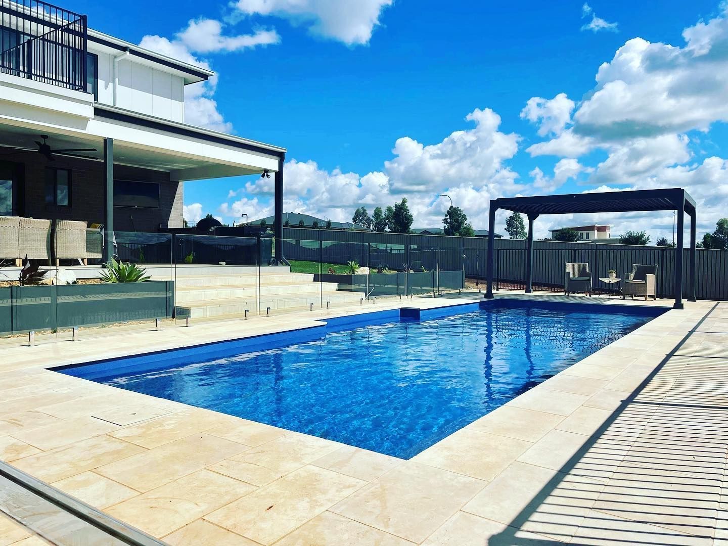 Blue-tiled pool with beige stone surround, pergola, and house under a bright blue sky.