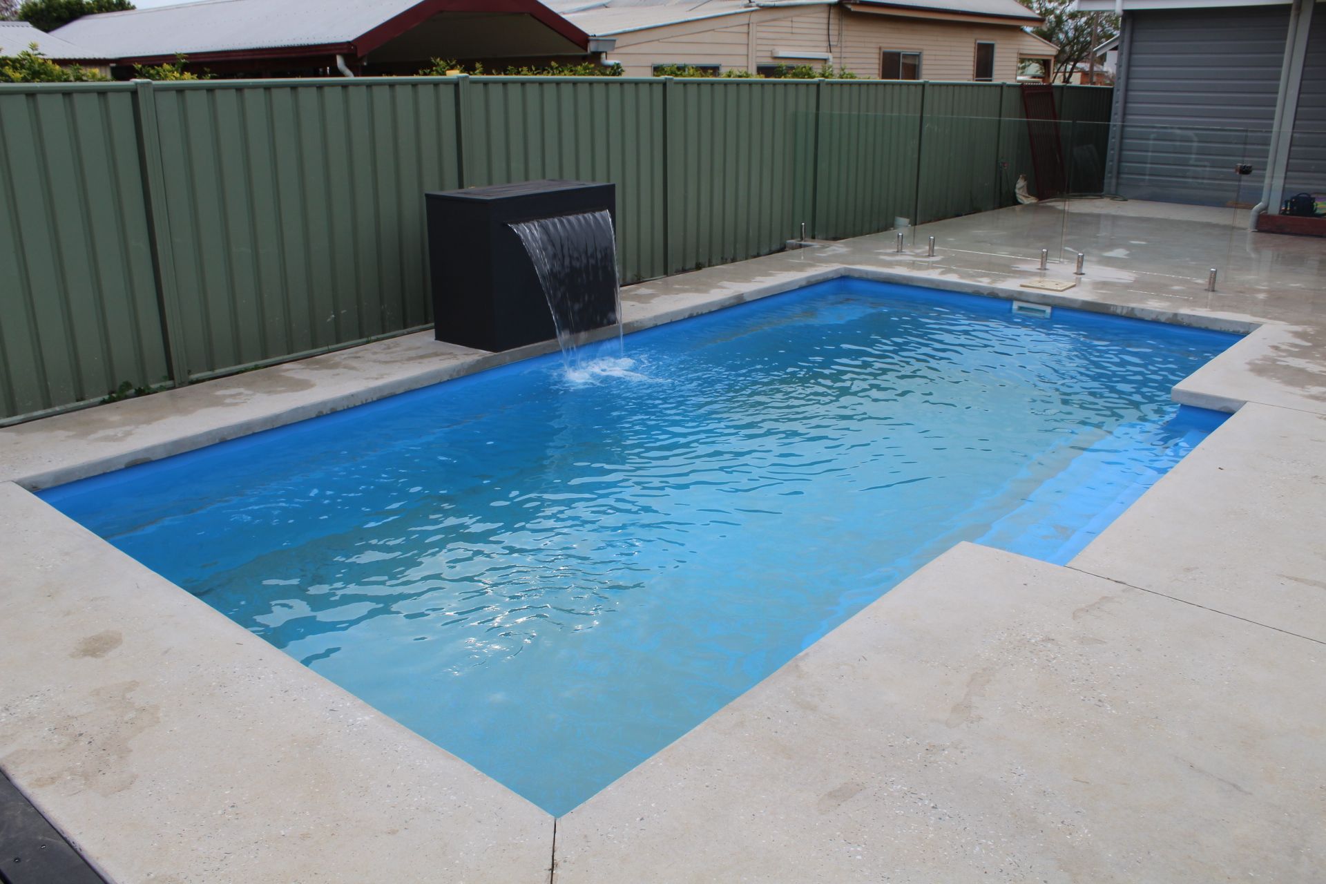 Rectangular swimming pool with blue water, concrete surround, and a black water feature against a green fence.