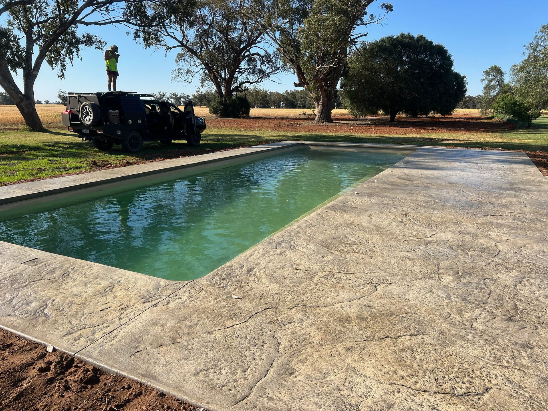 Swimming pool with textured concrete surround, green water, and a small vehicle on the grassy yard.