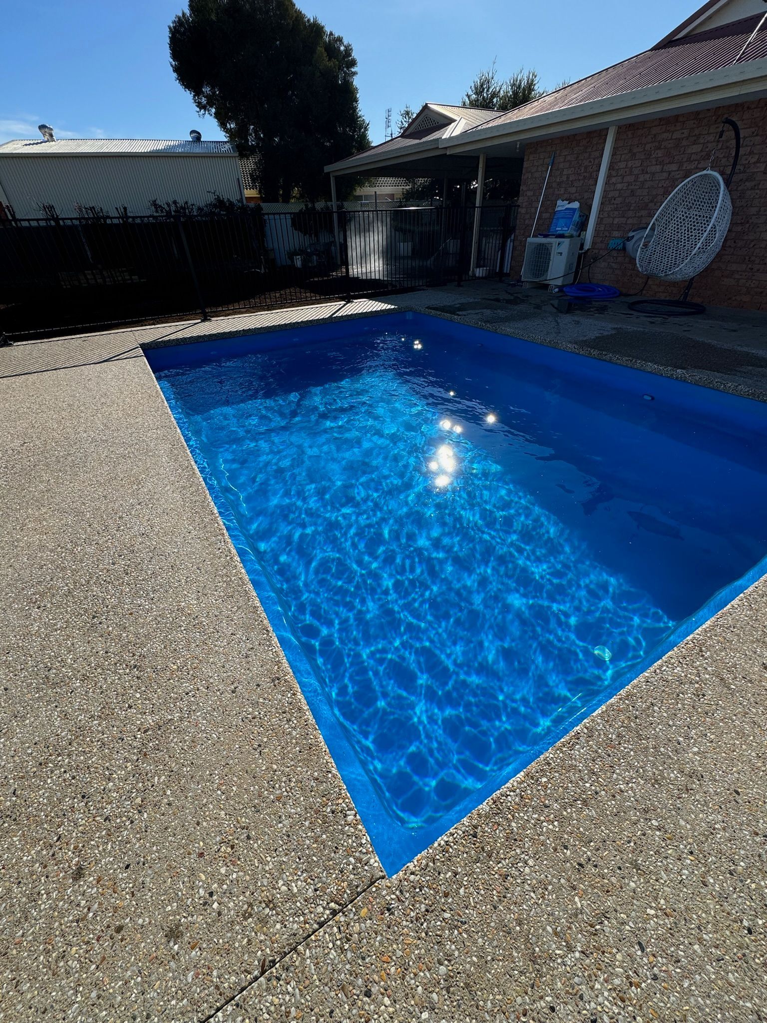 Blue rectangular swimming pool in backyard with pebbled concrete surround. Sunny day.