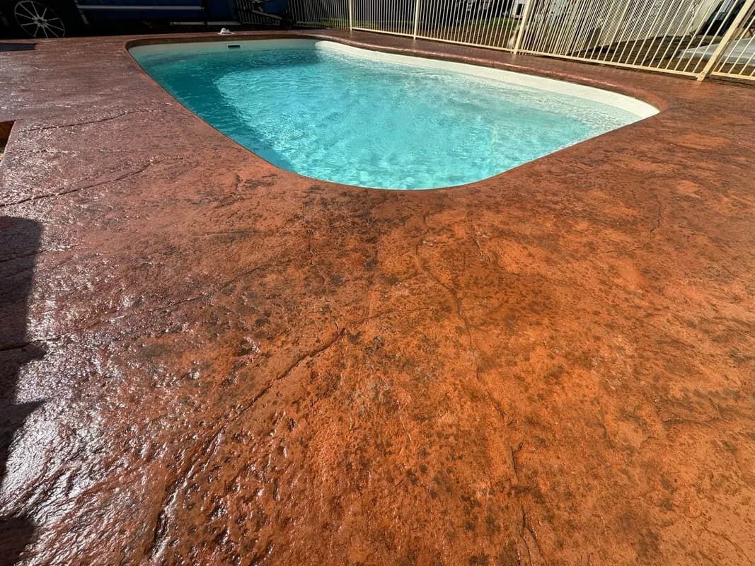 Pool with brown textured concrete deck. Blue water, sunny outdoor setting.