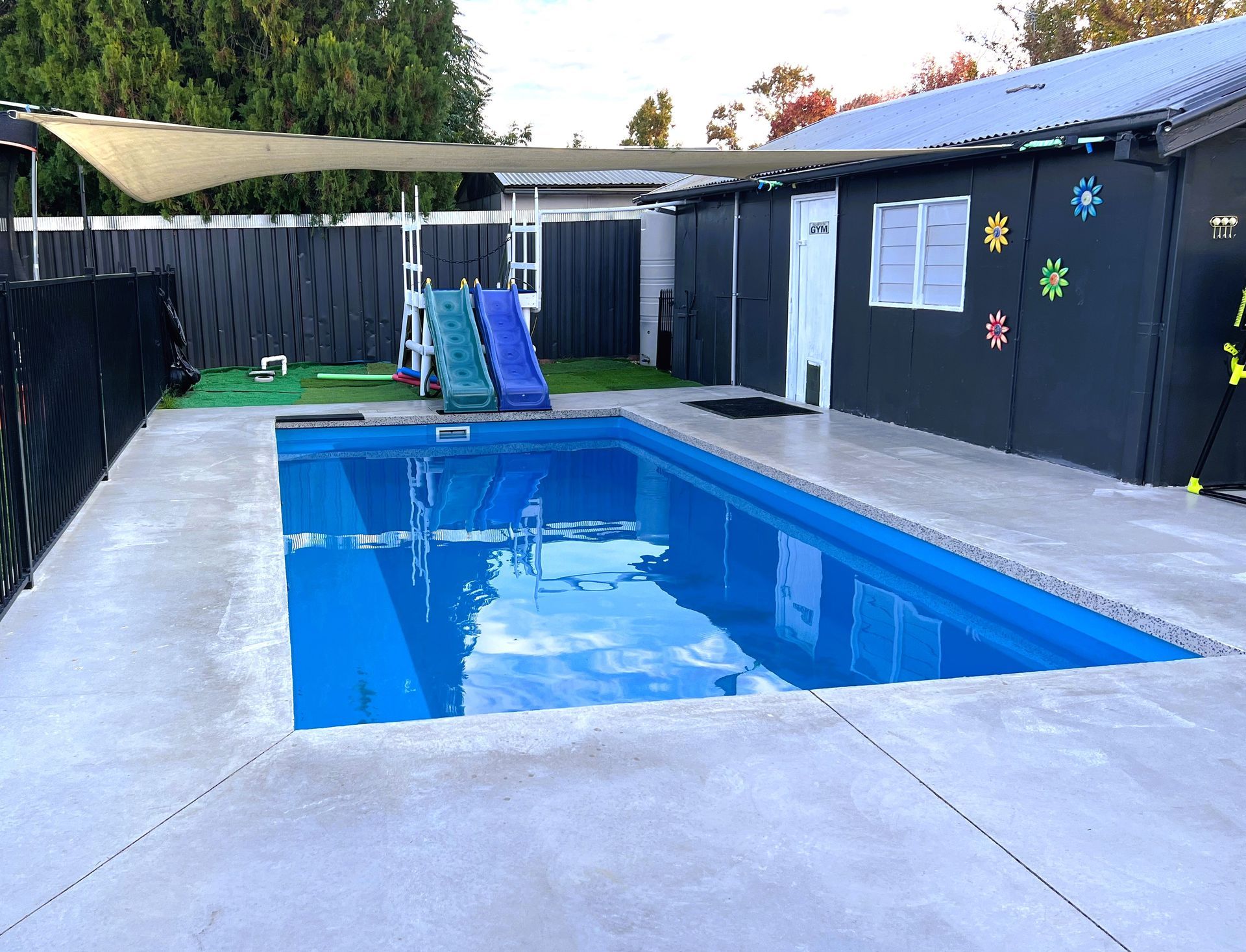 Backyard with rectangular pool, slide, and shade sail. Black fence and gray building with flowers.