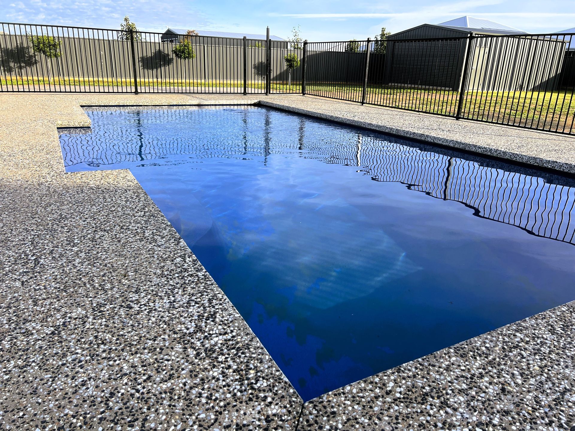 Rectangular swimming pool with gray pebble edging; blue water reflects sky. A black fence surrounds the yard.