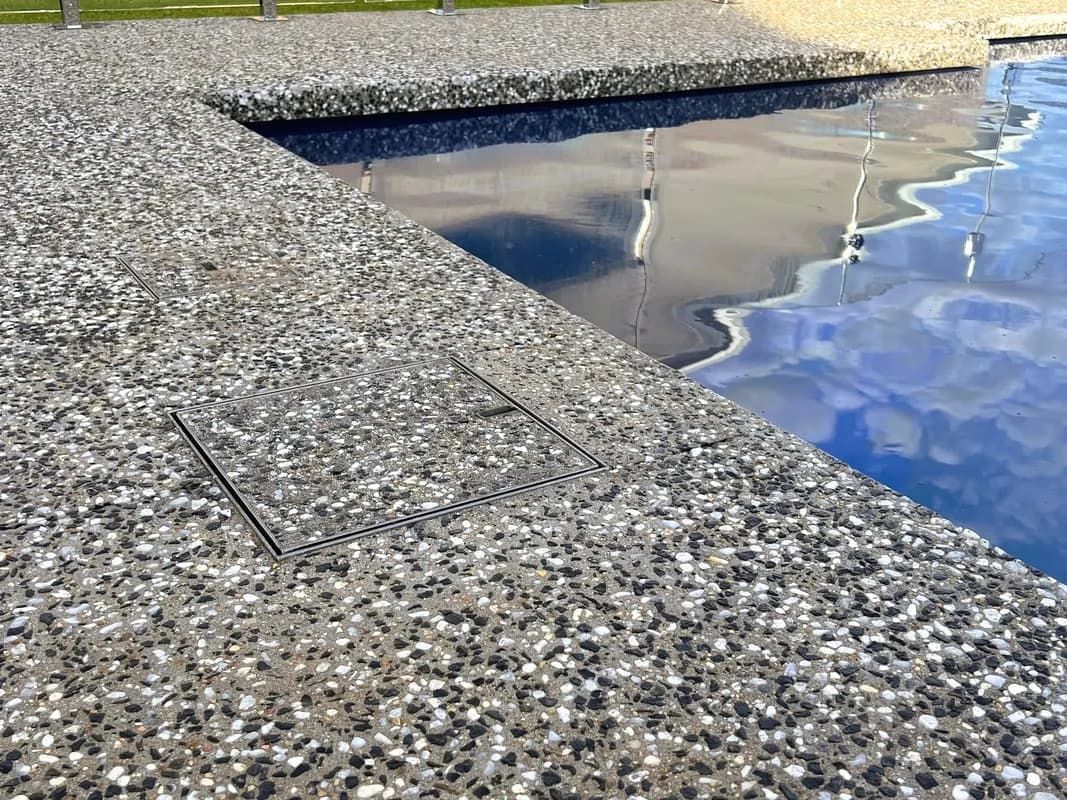 Close-up of a pool's edge with a speckled, gray concrete surface and a glimpse of the blue water.