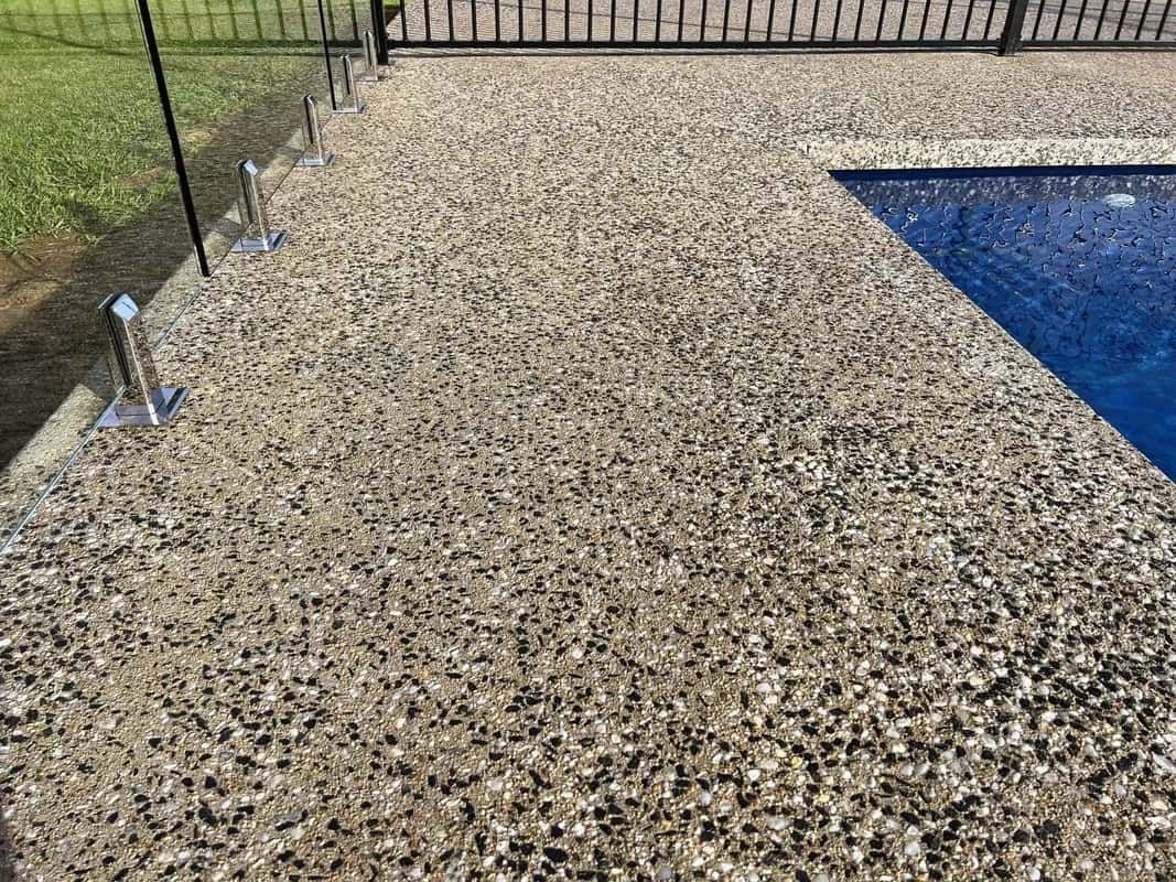 Concrete pool deck with exposed aggregate stones next to a blue pool and a black metal fence.
