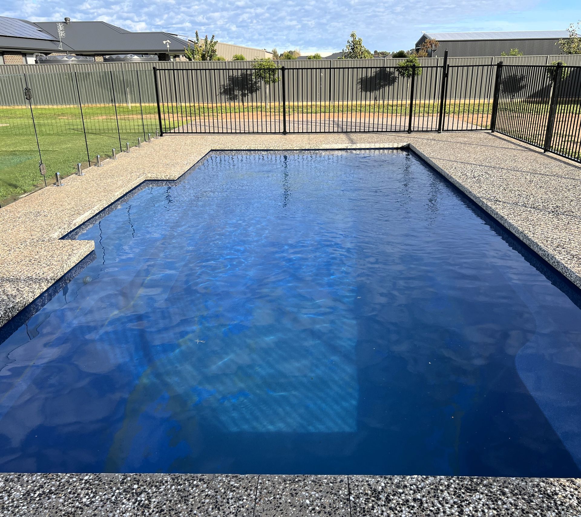 Rectangular swimming pool with blue water, surrounded by gray stone and a black fence, on a sunny day.