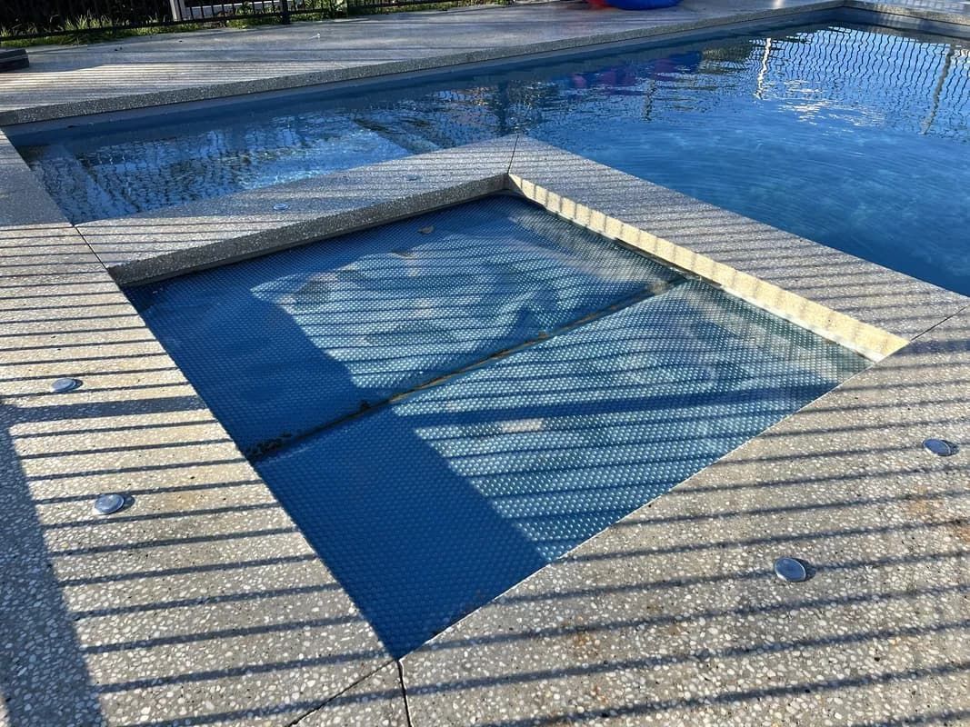 A rectangular pool with a built-in spa. The spa cover is partially open. Concrete decking surrounds the water.