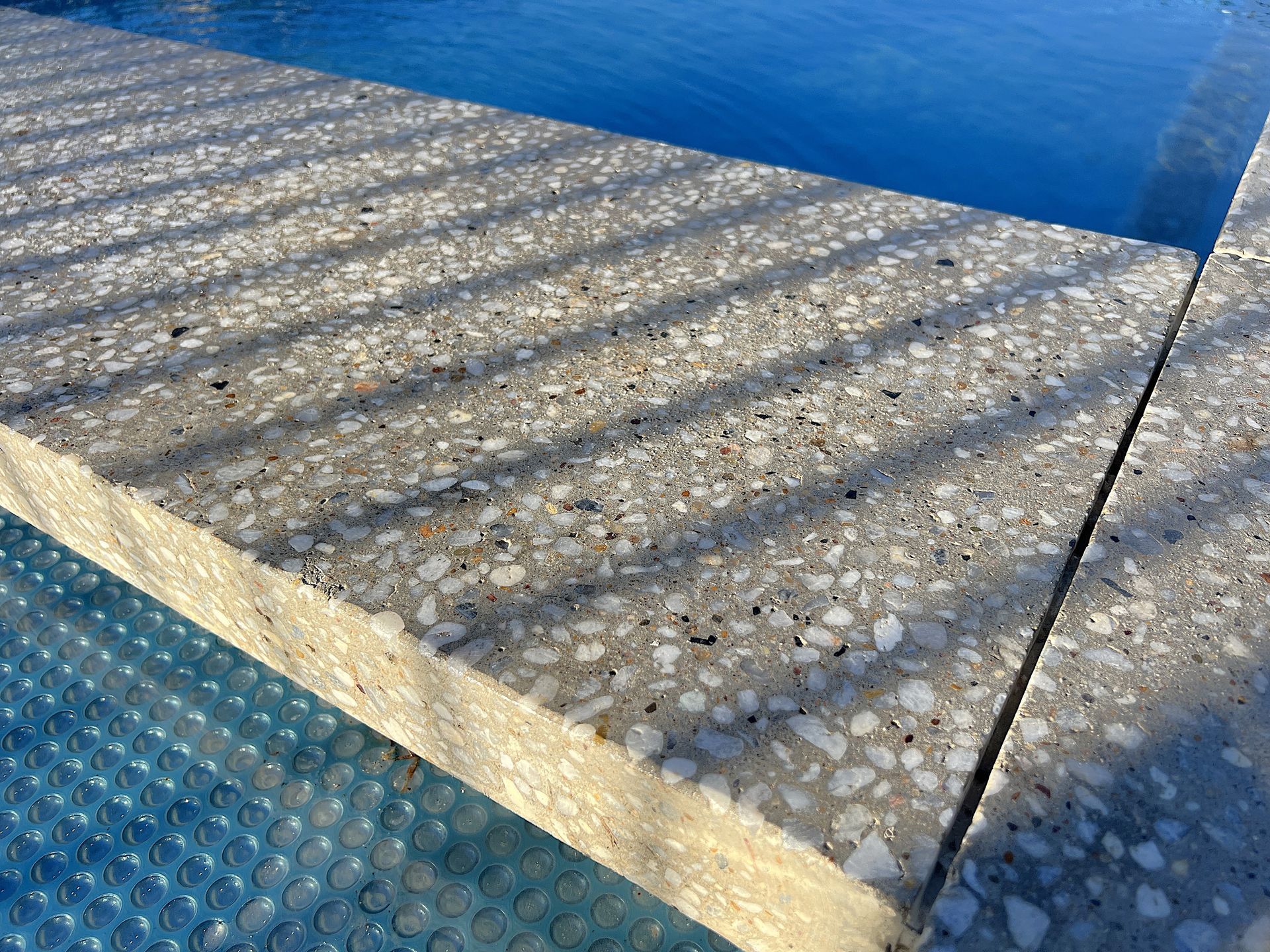 Poolside concrete with sunlight shadows cast on the surface, with pool water and blue cover visible.