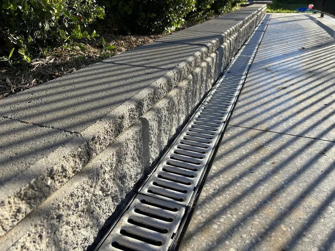 Concrete drainage channel with a metal grate, next to a concrete wall and pavement. Shadows cast across the surfaces.