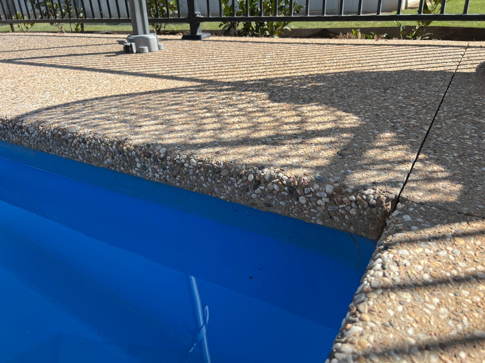Pool edge with exposed aggregate concrete coping and blue water. Shadow of fence on deck.