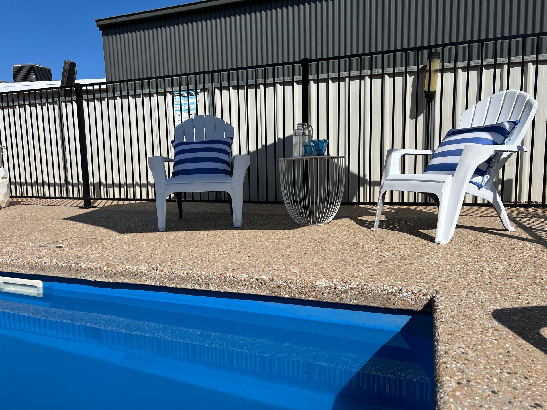 Two white chairs with blue striped pillows by a pool. Silver table, black fence, and building in the background.