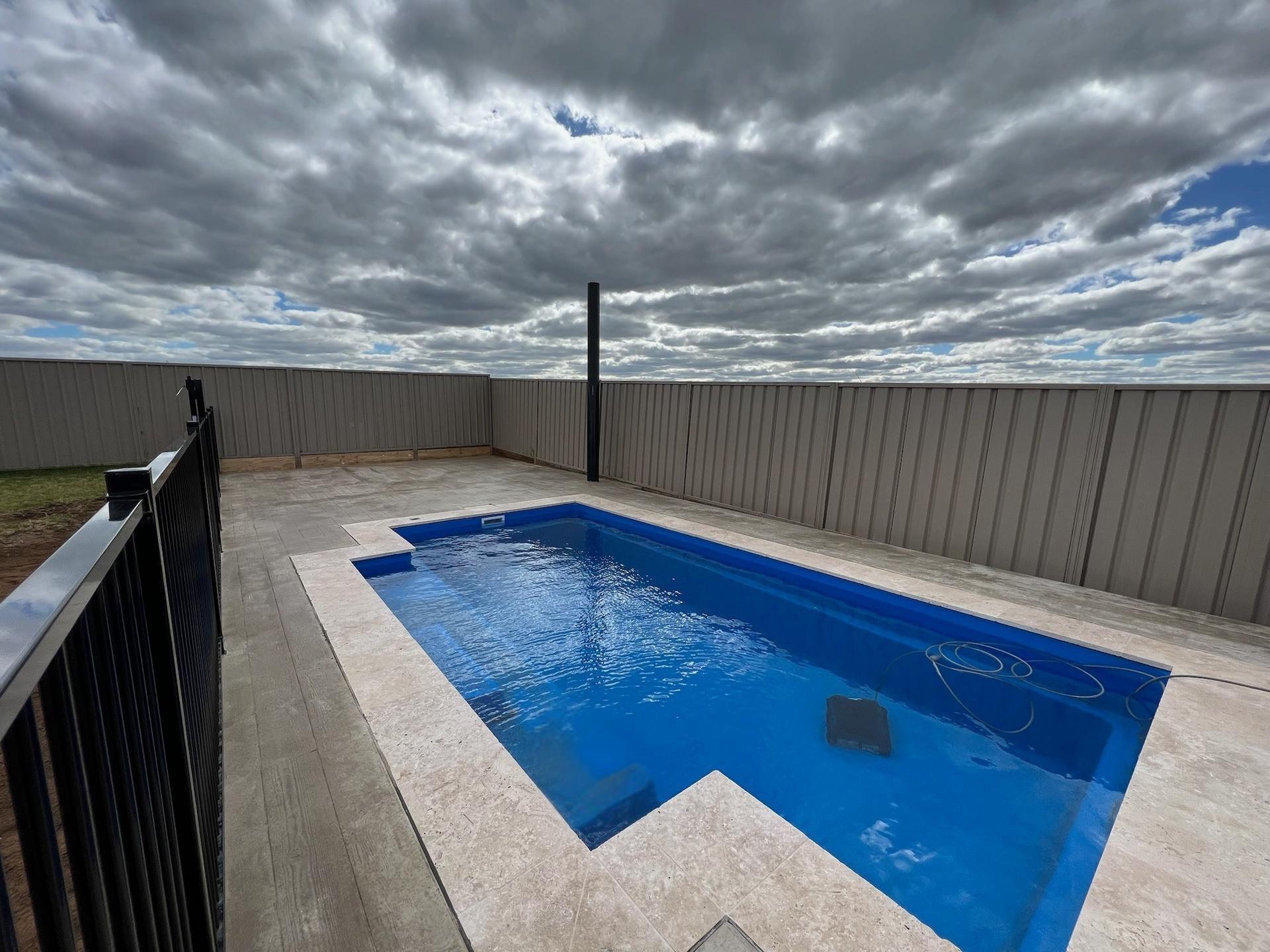 Small rectangular blue swimming pool surrounded by concrete and a high fence under a cloudy sky.
