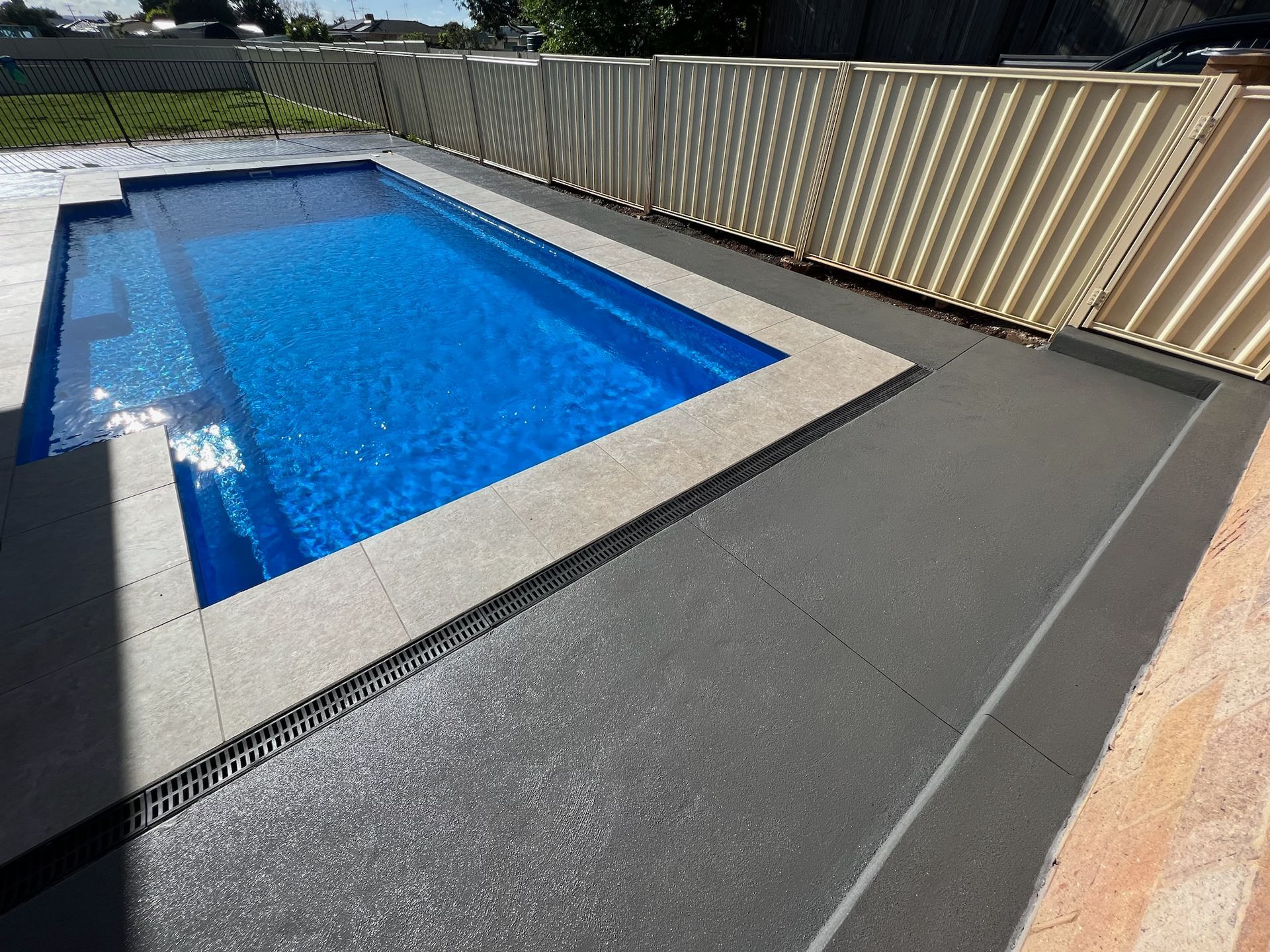 Rectangular swimming pool with blue water, surrounded by gray concrete and beige fence. Sunny outdoor setting.