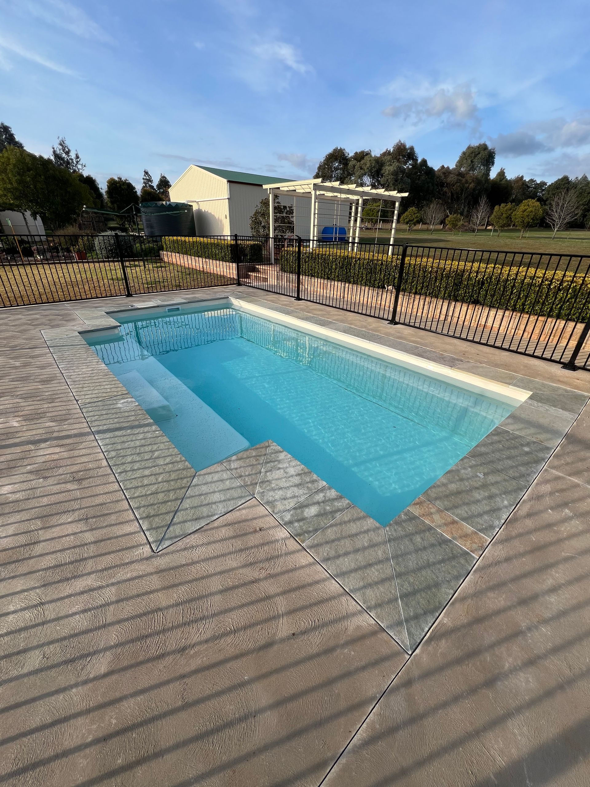 Rectangular pool with steps, surrounded by stone paving and a black fence, with a white building in the background.