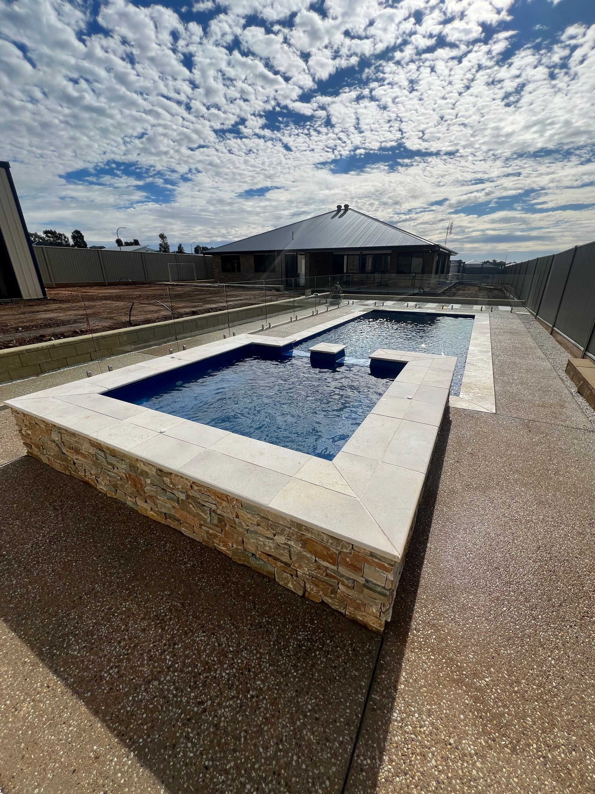 Swimming pool with blue tiles, stone border, and gravel surrounding. Cloudy sky.