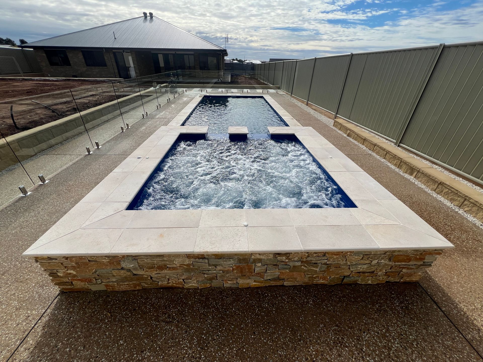 Rectangular jacuzzi with bubbly water, surrounded by stone edging and concrete patio, in front of a house.