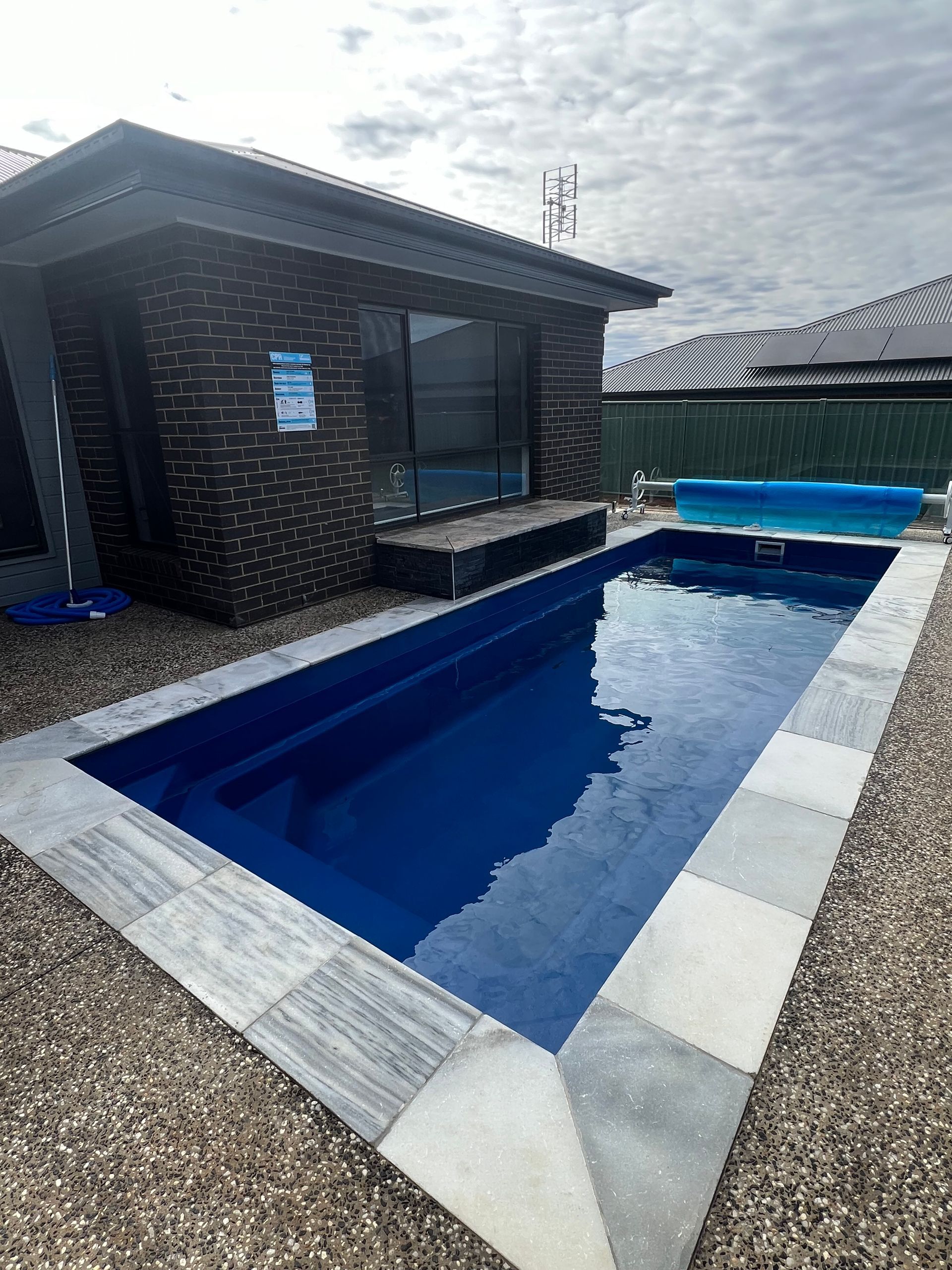 Rectangular blue pool with grey stone surround next to a brick building.